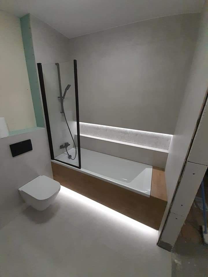 Nowoczesna łazienka w trakcie wykańczania: biała wanna z czarną kabiną prysznicową, podwieszana toaleta, oświetlenie LED pod wanną i wnęką, surowe ściany w szarym odcieniu, widoczne elementy...