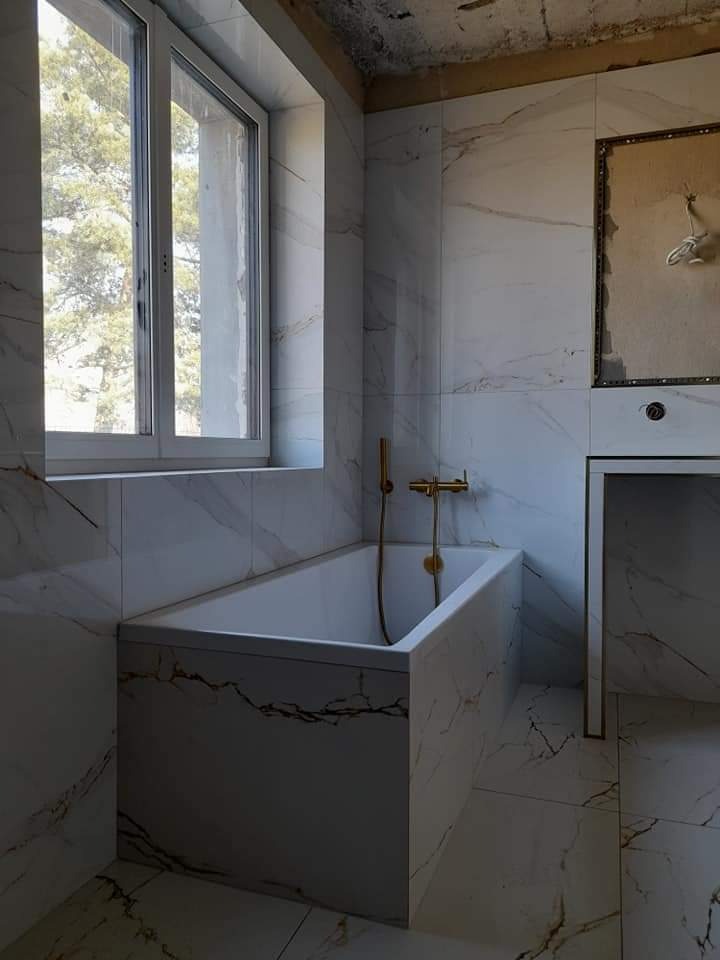 Wanna obudowana płytkami imitującymi marmur z złotymi żyłami, bateria w kolorze złota, okno z widokiem na drzewo, surowy sufit w trakcie prac wykończeniowych.