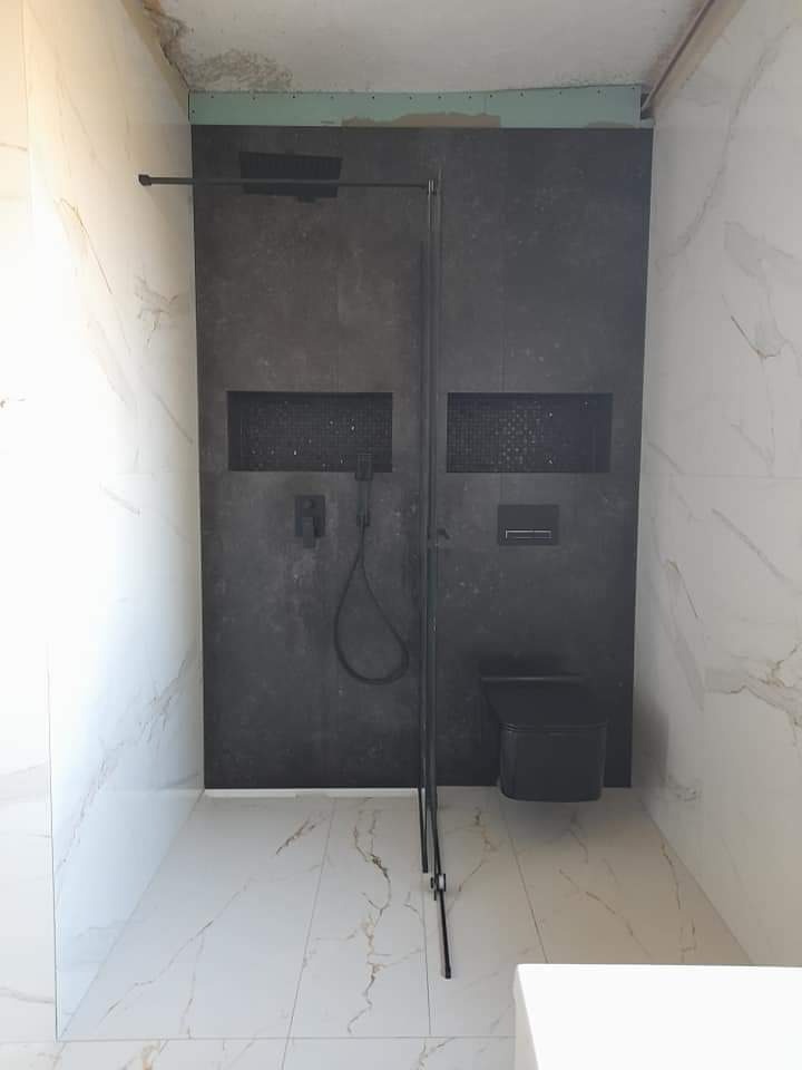 Wnętrze łazienki w trakcie remontu: białe marmurowe płytki kontrastują z czarną ścianą prysznicową z wnękami, widoczna czarna armatura i toaleta, surowy sufit z zieloną płytą gipsową.