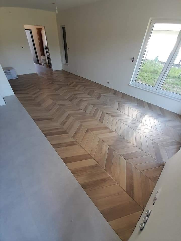 Ułożona w jodełkę podłoga z drewnopodobnych płytek w jasnym, przestronnym wnętrzu. Widoczne duże okno z widokiem na zieleń i fragmenty surowych ścian przed wykończeniem.