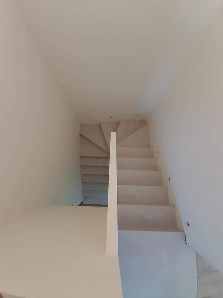 Nowoczesne, minimalistyczne schody w jasnej tonacji, prowadzące w górę, z oświetleniem punktowym na ścianie.