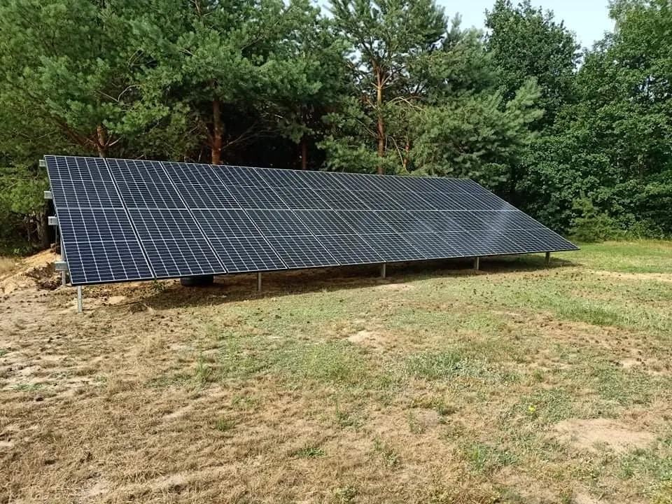Panel słoneczny ustawiony na metalowych podporach na tle lasu sosnowego, widoczne ślady po pracach ziemnych.