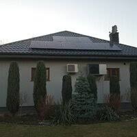 Dom jednorodzinny z panelami słonecznymi na dachu i klimatyzatorem na ścianie, otoczony iglakami i krzewami.