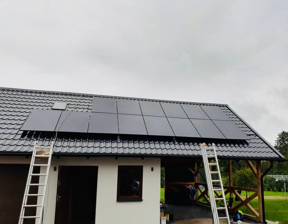 Instalacja paneli fotowoltaicznych na dachu domu z dachówką, widoczne dwie aluminiowe drabiny opierające się o budynek.