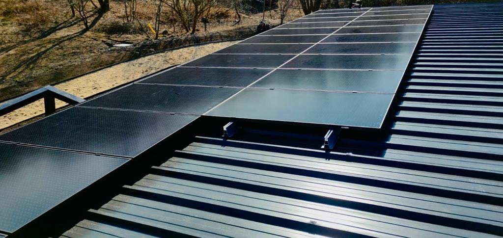 Montaż paneli słonecznych na metalowym dachu z widocznymi elementami mocującymi i fragmentem otoczenia z drzewami w tle.