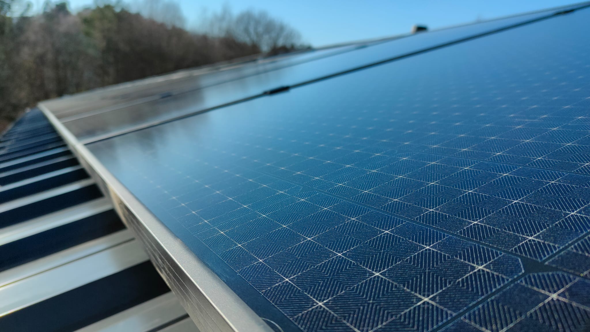 Błękitny panel słoneczny z wyraźną, geometryczną strukturą na dachu z blachy trapezowej, w tle drzewa i jasne niebo.