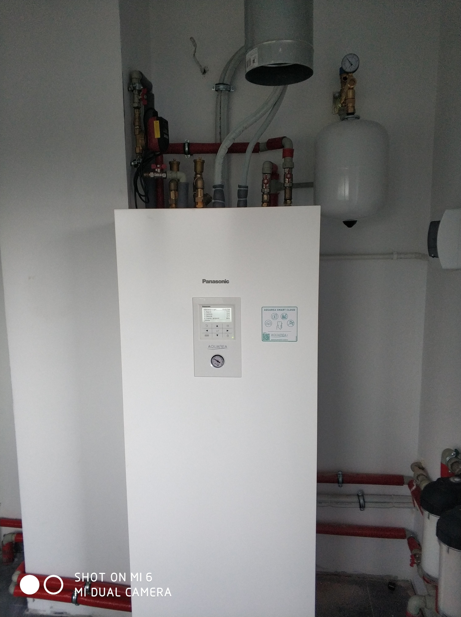 Biała pompa ciepła Panasonic Aquarea w kotłowni w Brzostku, z widocznymi rurami instalacji grzewczej i zbiornikiem wyrównawczym.