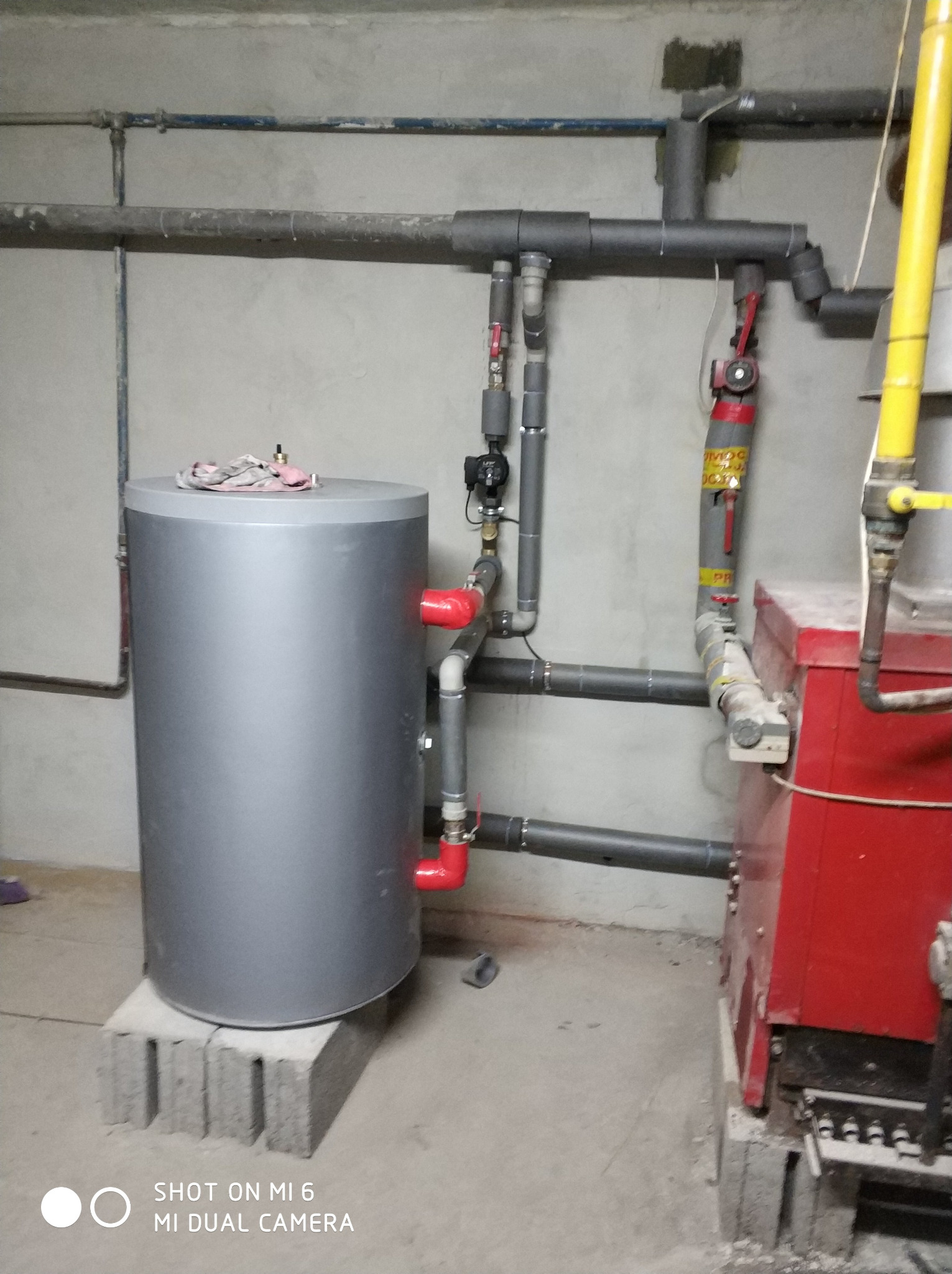 Szary zbiornik akumulacyjny ciepła, umieszczony na betonowych bloczkach, z podłączonymi rurami z czerwonymi złączkami, obok czerwonego kotła i szarej instalacji rurowej na tle surowej ściany...