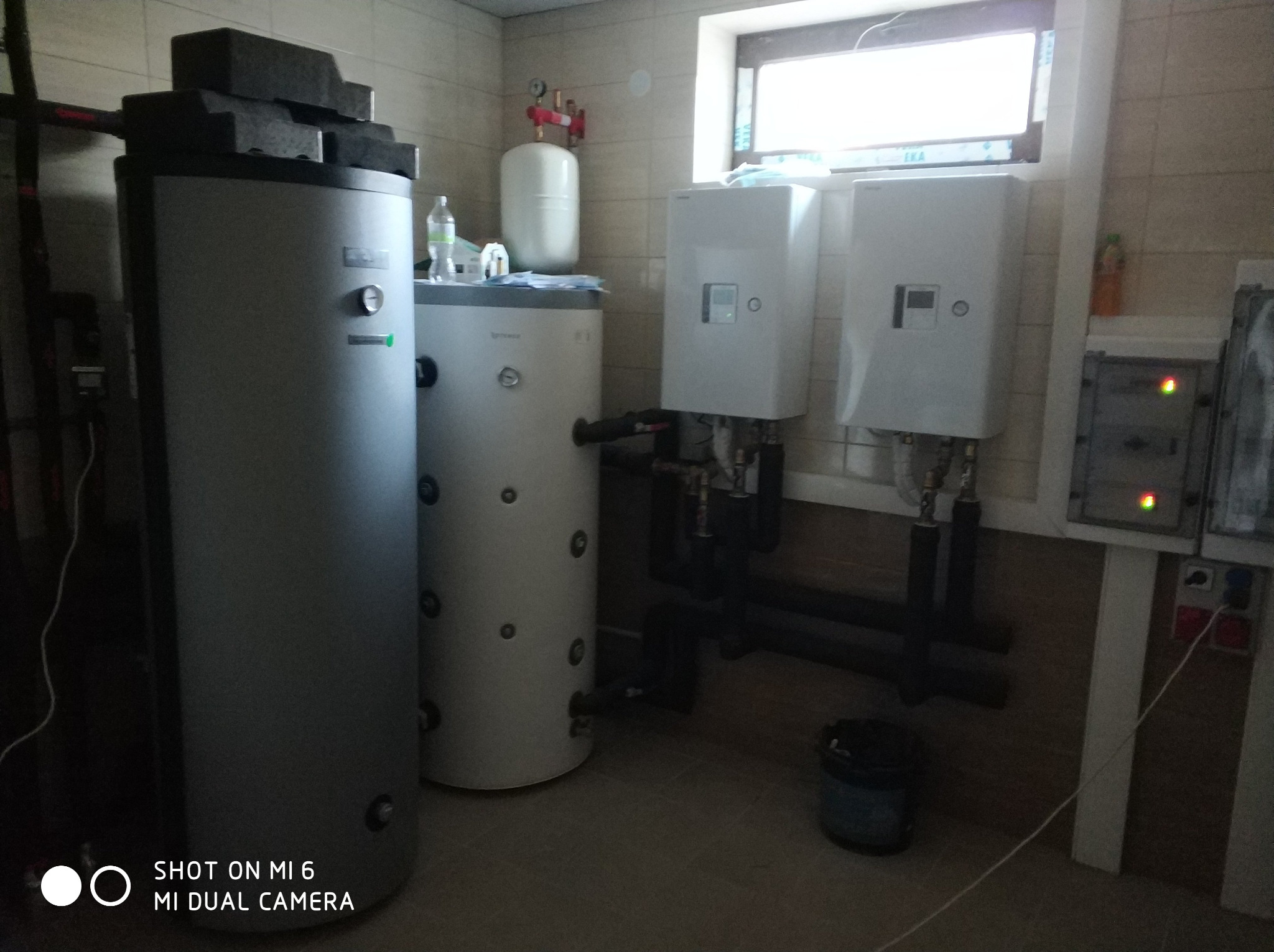 Dwa białe zbiorniki akumulacyjne ciepła stoją obok dwóch białych jednostek wewnętrznych, z widocznymi rurami i osprzętem, zainstalowane w pomieszczeniu z beżowymi płytkami, pod oknem.