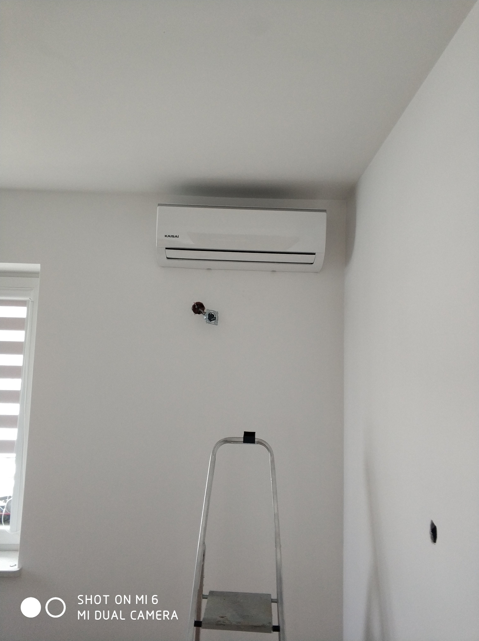 Biały klimatyzator Kaisai zamontowany na jasnej ścianie nad metalową drabiną, widoczne przygotowane gniazdka elektryczne.