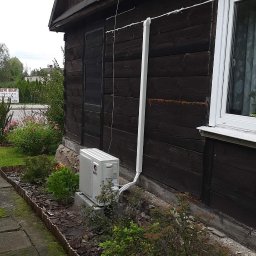 Bartłomiej Bieliński - Zewnętrzna jednostka klimatyzacji marki Gree zamontowana przy ciemnej, drewnianej ścianie domu z widocznymi rurami odprowadzającymi skropliny, otoczona rabatą z roślinami i kamienną ścieżką.