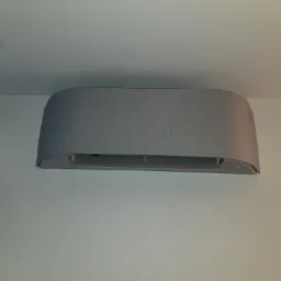 Szara, nowoczesna klimatyzacja ścienna z delikatną teksturą, zamontowana w jasnym wnętrzu, widoczny fragment okna z błękitnym budynkiem w tle.