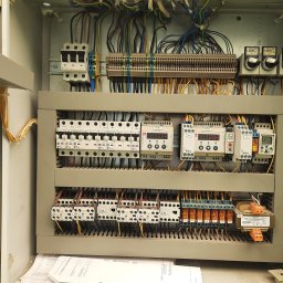 Bartłomiej Bieliński - Otwarta szafa sterownicza z widocznymi bezpiecznikami, przekaźnikami, przewodami i schematami elektrycznymi, zainstalowana wewnątrz pomieszczenia.