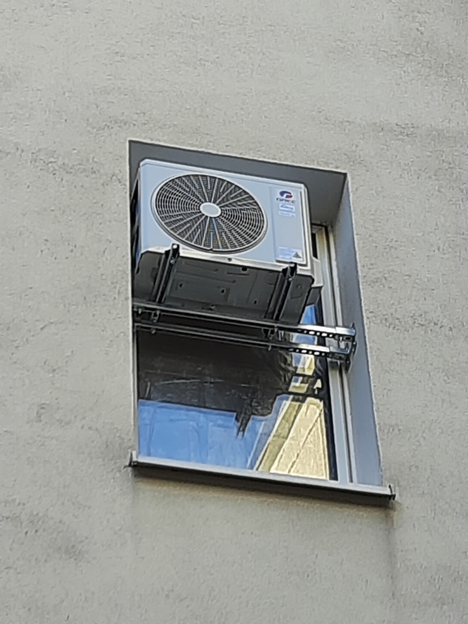 Jednostka zewnętrzna klimatyzatora zamontowana w oknie budynku, widoczna z zewnątrz, na specjalnym metalowym stelażu.