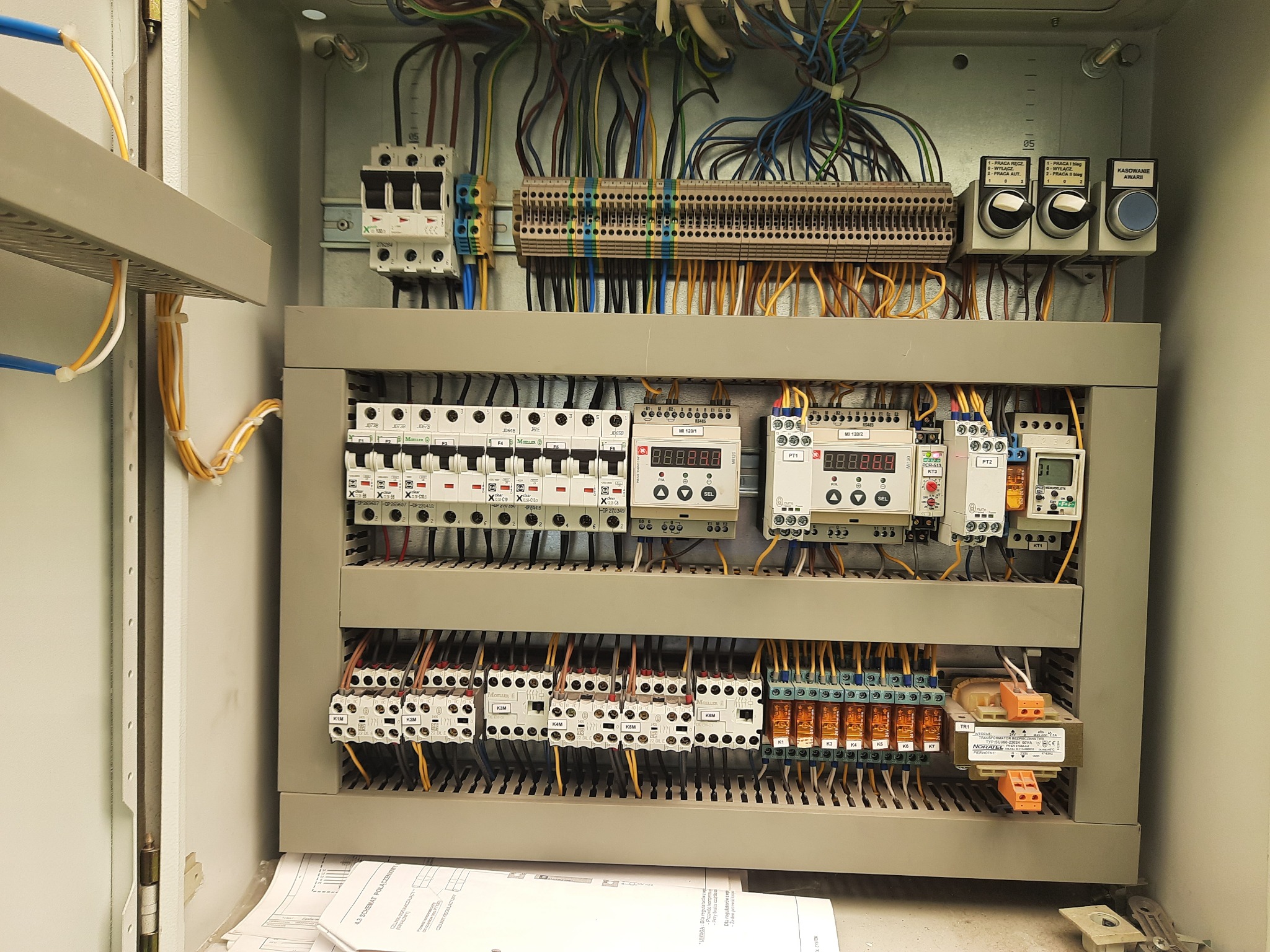 Otwarta szafa sterownicza z widocznymi bezpiecznikami, przekaźnikami, przewodami i schematami elektrycznymi, zainstalowana wewnątrz pomieszczenia.