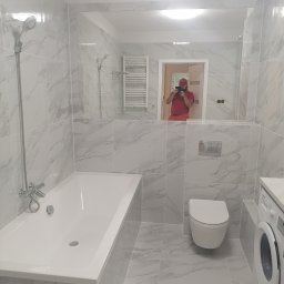 Adam Macina FACHURA - Wykończona łazienka z biało-szarymi marmurkowymi płytkami, białą wanną, toaletą i pralką; w lustrze odbija się osoba robiąca zdjęcie.