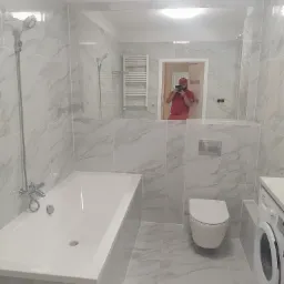 Wykończona łazienka z biało-szarymi marmurkowymi płytkami, białą wanną, toaletą i pralką; w lustrze odbija się osoba robiąca zdjęcie.