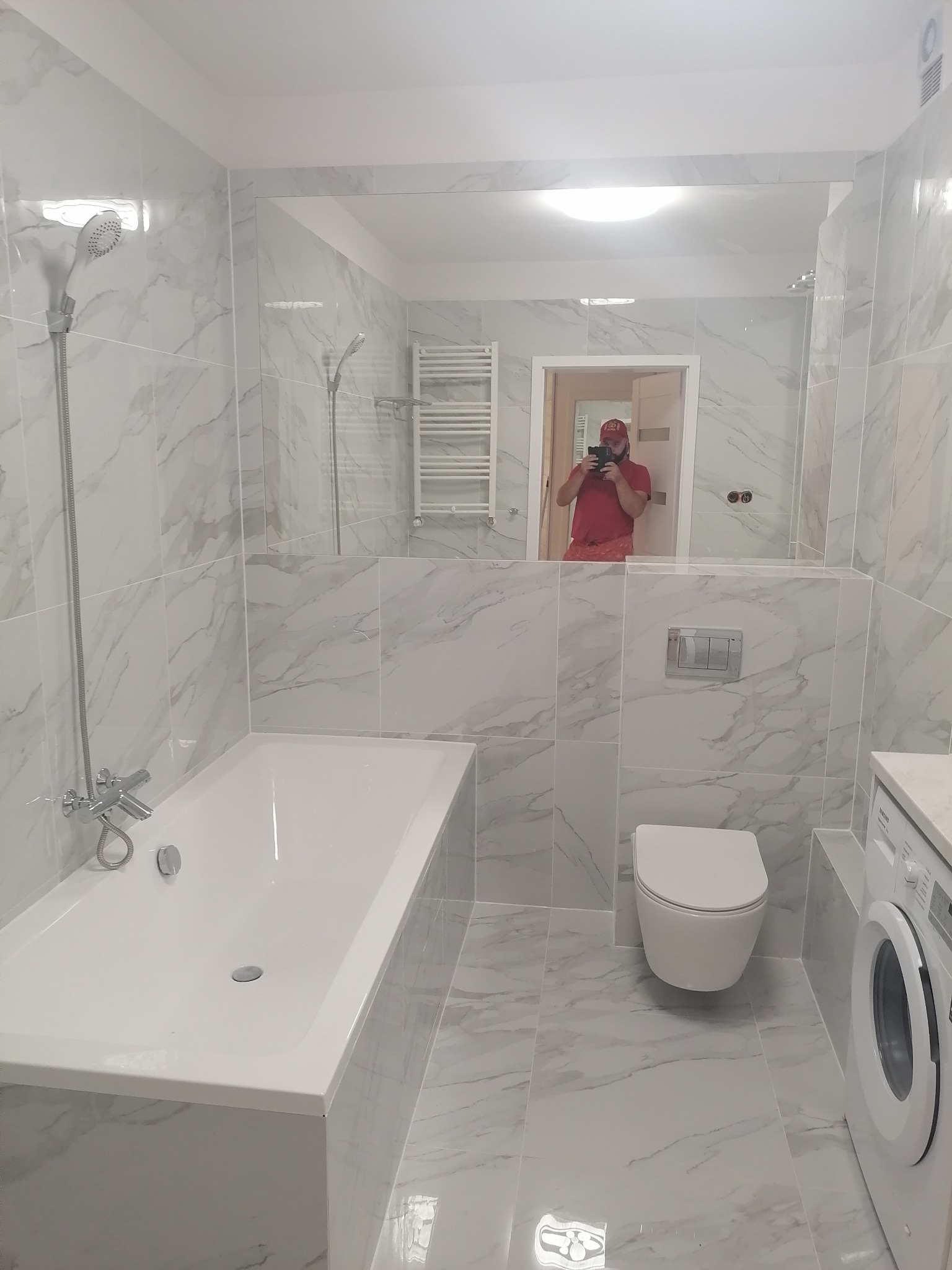 Wykończona łazienka z biało-szarymi marmurkowymi płytkami, białą wanną, toaletą i pralką; w lustrze odbija się osoba robiąca zdjęcie.