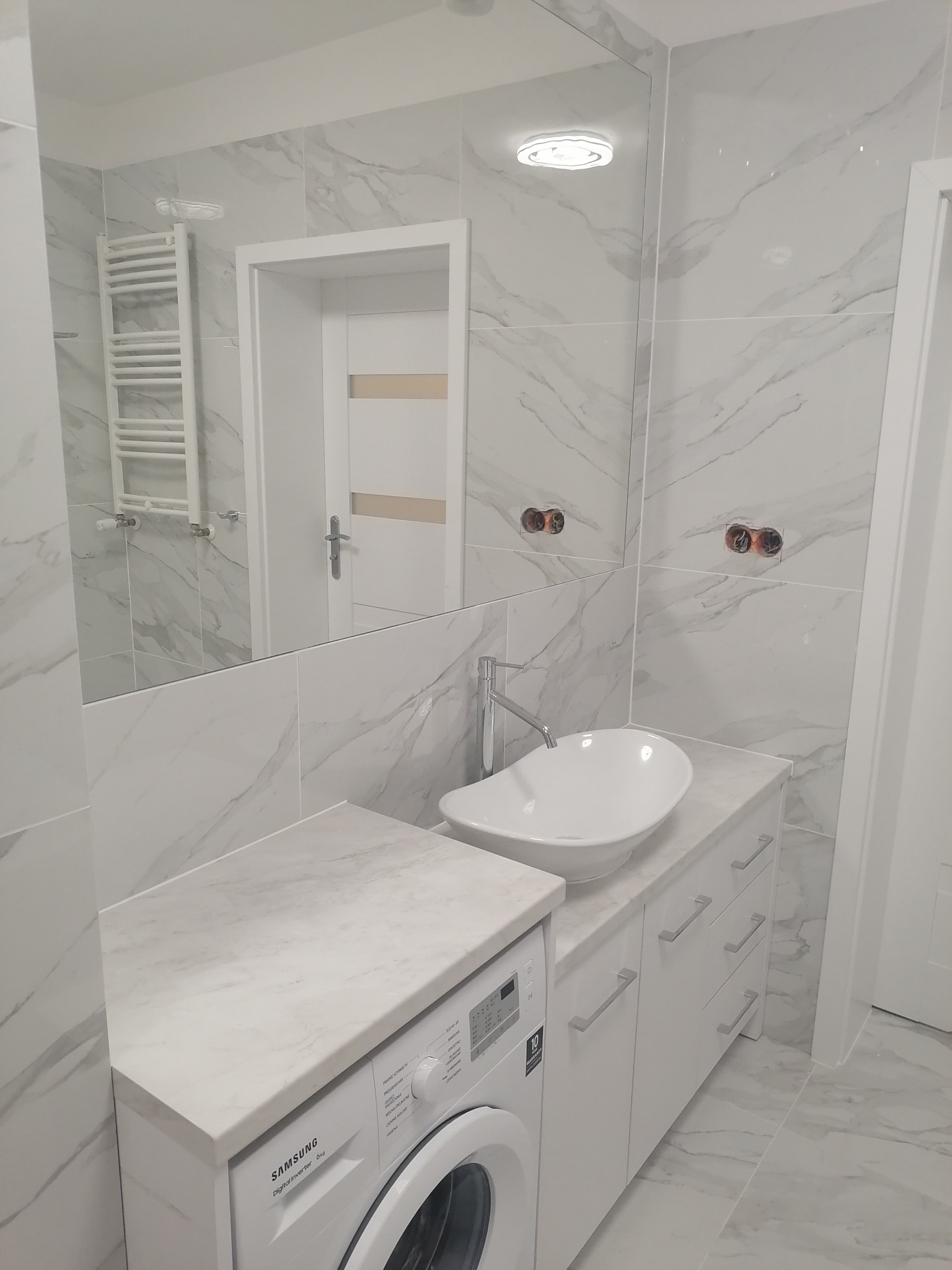 Nowoczesna łazienka z marmurowymi płytkami, umywalką nablatową, białą szafką pod umywalką, pralką pod blatem i grzejnikiem drabinkowym widocznym w lustrze.