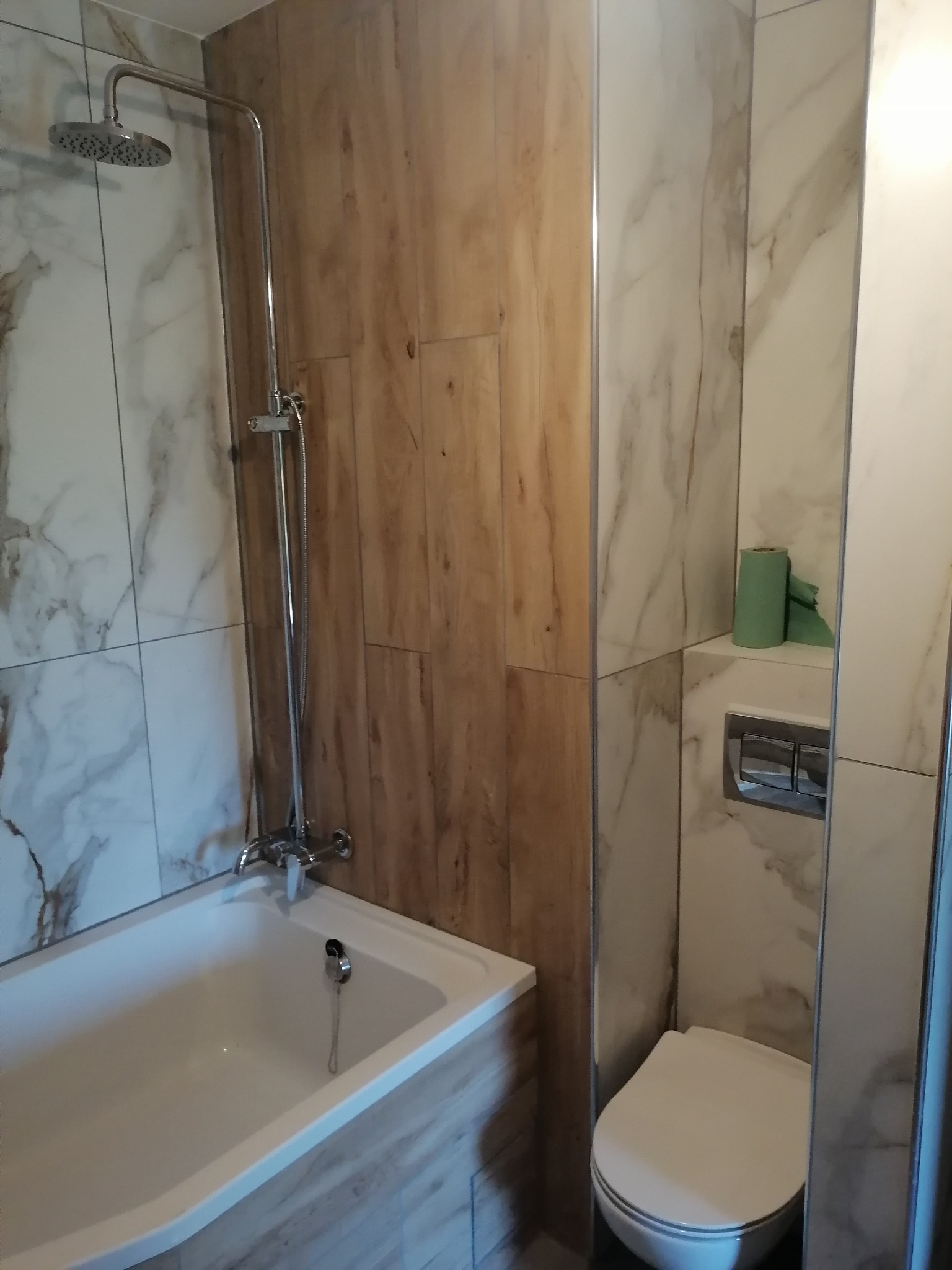 Wykończona łazienka z wanną, prysznicem i toaletą, wyłożona płytkami imitującymi drewno i marmur, z rolką zielonego papieru toaletowego.