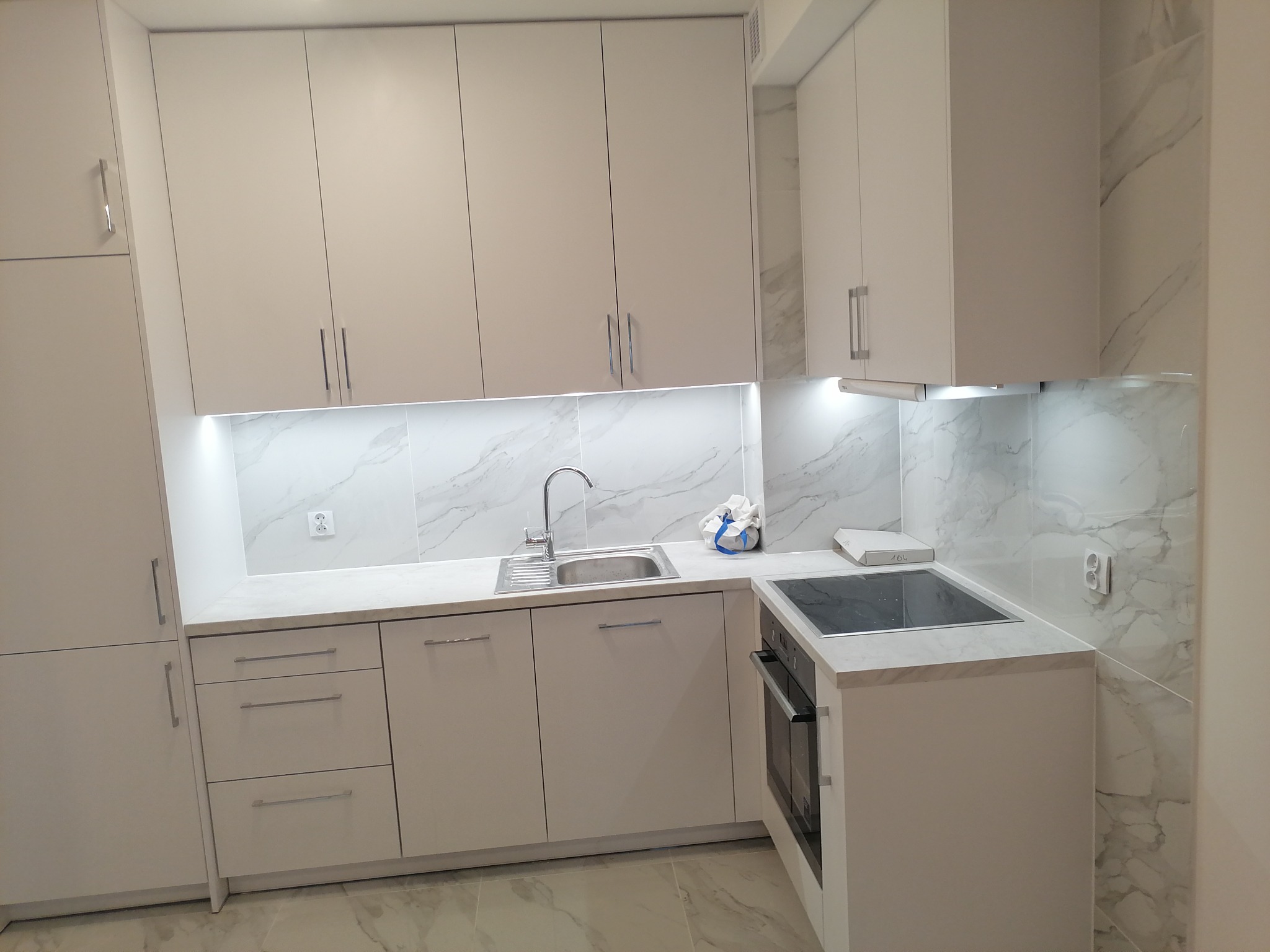 Nowoczesna, jasna kuchnia z białymi szafkami, marmurowym blatem i backsplashiem, oświetleniem LED pod szafkami wiszącymi i płytą indukcyjną.