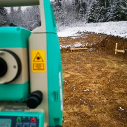 Zielono-beżowy teodolit ustawiony na wykopie pod fundamenty, w tle ośnieżony las.