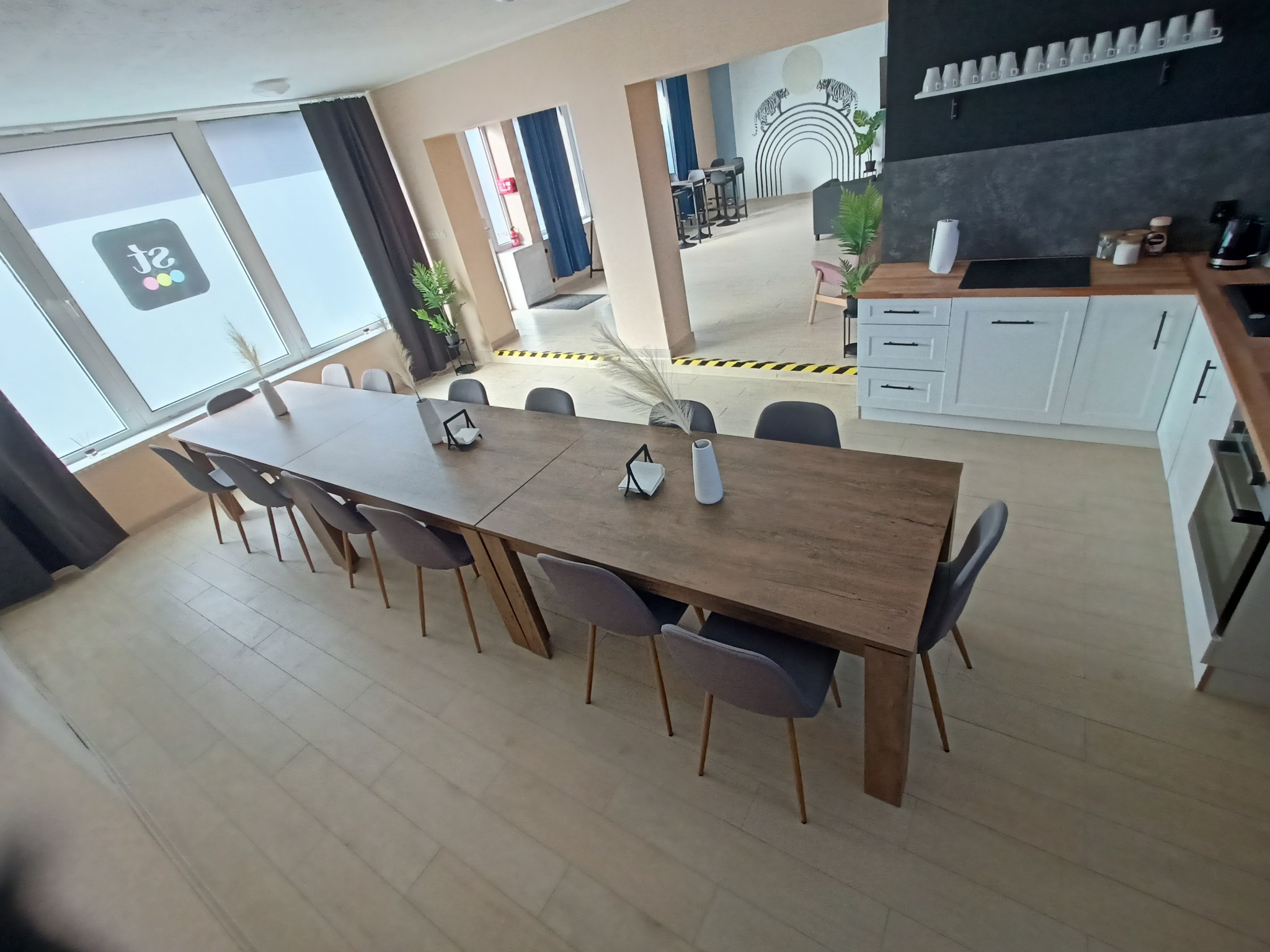 Wnętrze sali konferencyjnej z długim drewnianym stołem otoczonym szarymi krzesłami, minimalistyczne dekoracje na stole, widok na kuchnię i dalszą część pomieszczenia z dekoracyjną ścianą w paski...