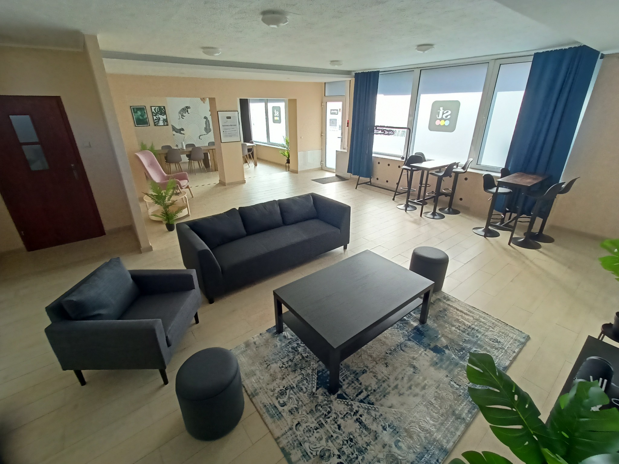 Przestronne wnętrze biurowe z szarą sofą, fotelem, stolikiem kawowym na wzorzystym dywanie, wysokimi stolikami barowymi przy oknach i roślinami doniczkowymi.