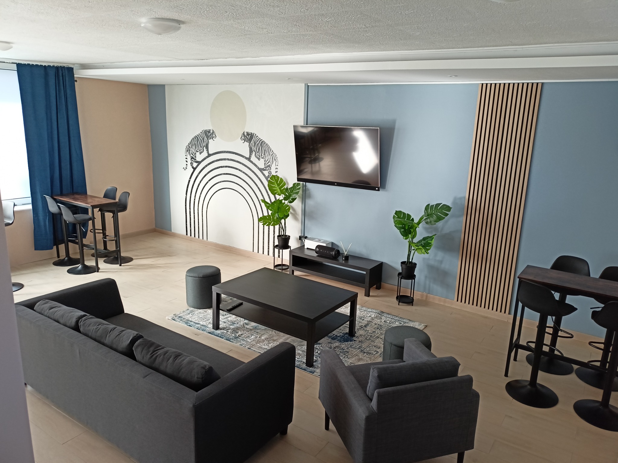 Nowoczesne wnętrze pokoju dziennego z szarą sofą, stolikiem kawowym, telewizorem, dekoracją ścienną z motywem tygrysów i tęczy oraz wysokimi stołkami barowymi.