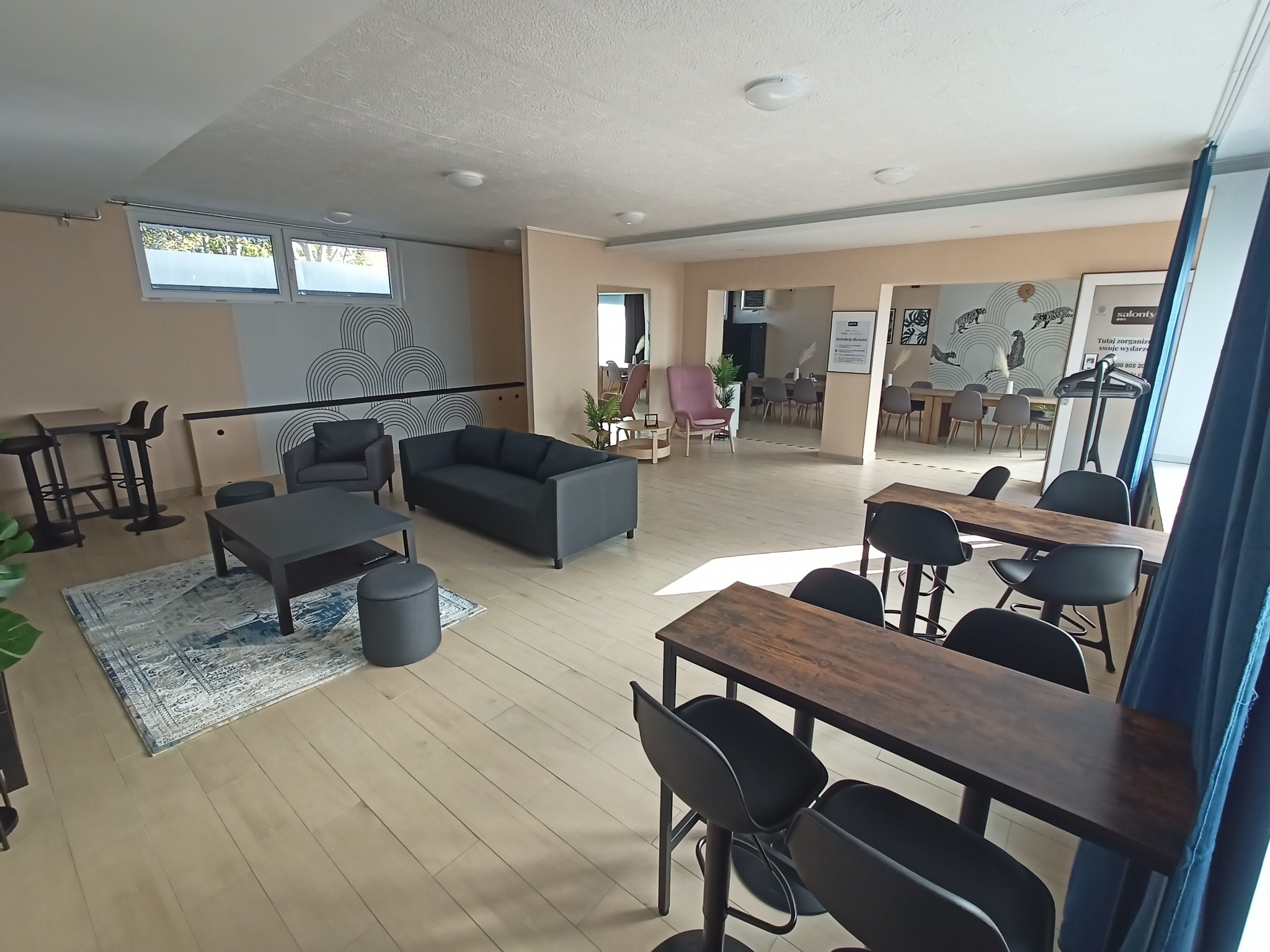 Przestronne wnętrze z szarymi meblami wypoczynkowymi, drewnianymi stołami barowymi i czarnymi krzesłami hokerami, udekorowane geometrycznym wzorem na ścianie i plakatem z motywami roślinnymi.