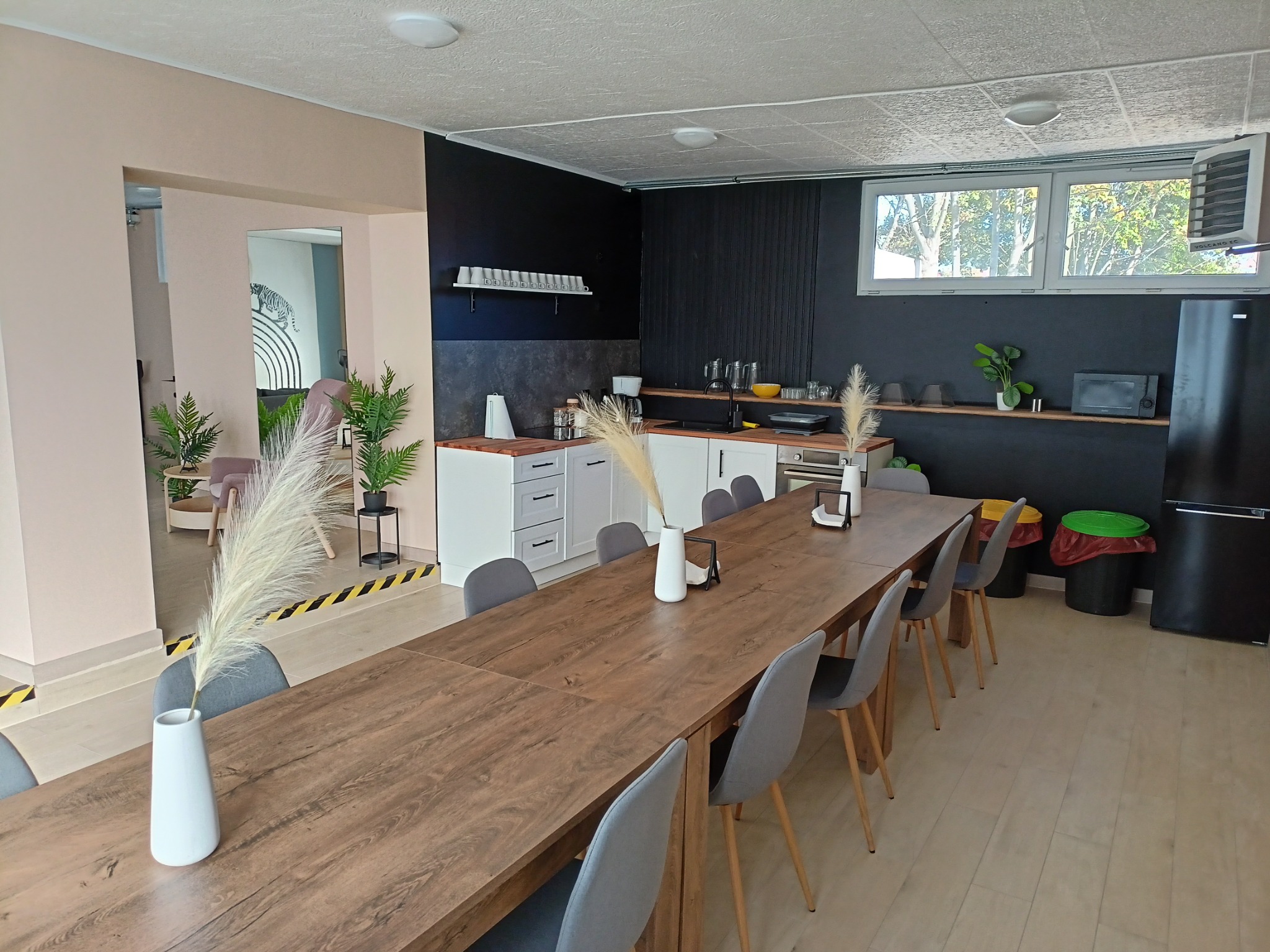 Sala konferencyjna z długim drewnianym stołem, szarymi krzesłami, dekoracjami z trawy pampasowej w wazonach, aneksem kuchennym w tle i widokiem na ogród przez okno.