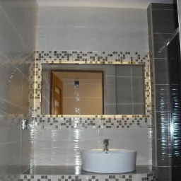 Nowoczesna łazienka z umywalką nablatową, lustrem z podświetleniem LED obramowanym mozaiką i szarymi płytkami imitującymi falowanie wody.