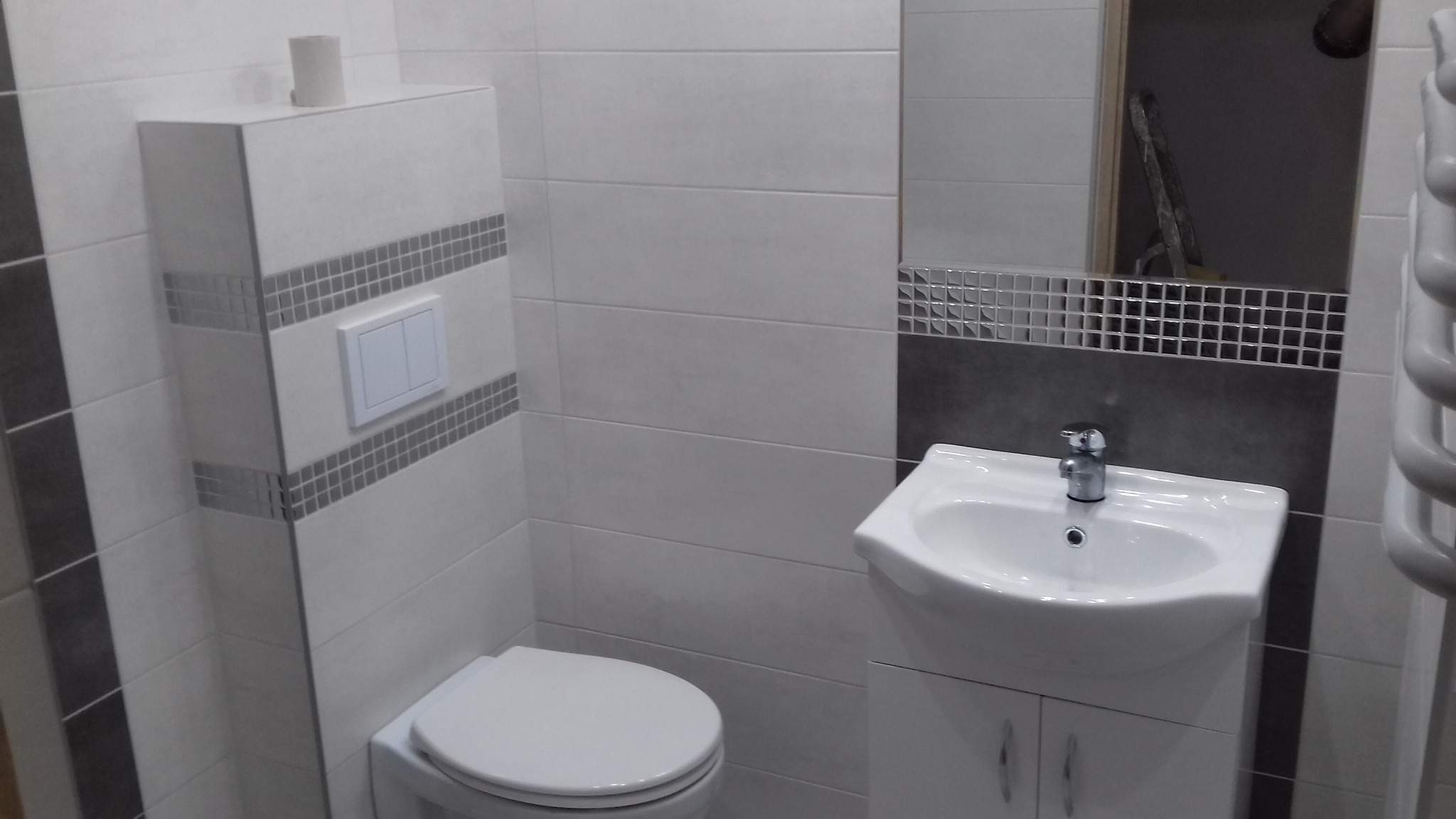 Nowoczesna łazienka z białymi płytkami, umywalką z szafką i podwieszaną toaletą z mozaikowymi akcentami, widoczne odbicie drabiny w lustrze.