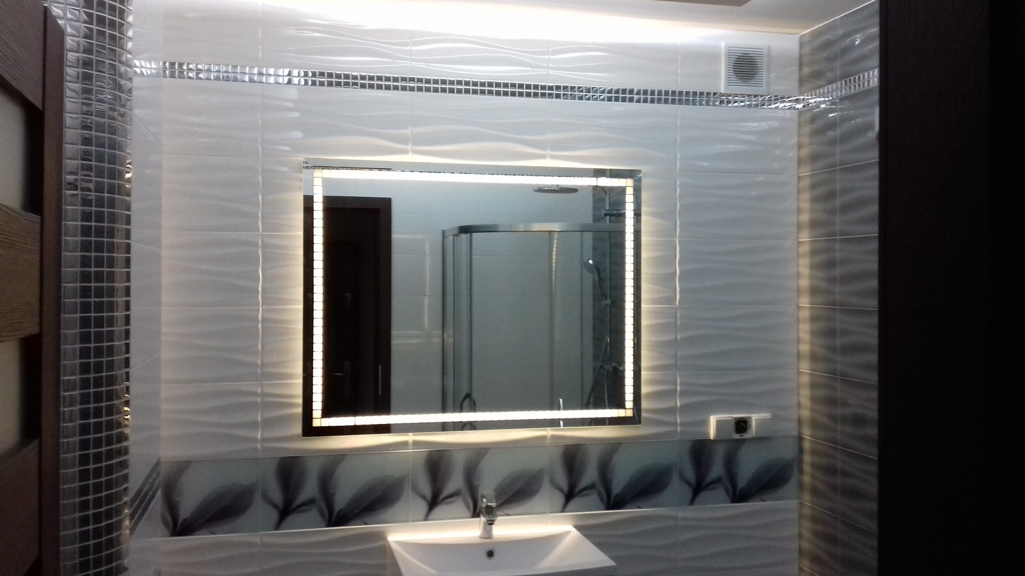 Nowoczesna łazienka z podświetlanym lustrem LED, umywalką i dekoracyjnymi płytkami ściennymi z motywem liści, widoczna kabina prysznicowa w odbiciu lustra.