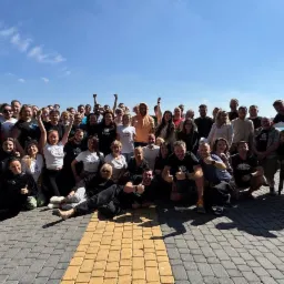 Duża grupa uśmiechniętych osób pozuje do zdjęcia na zewnątrz, część w koszulkach z logo, z widokiem na jezioro i budynek z trzcinowym dachem w tle.