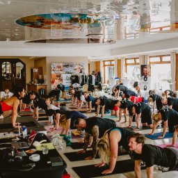 TIME4YOU Sp. z o.o. - Grupa osób ćwiczy jogę w jasnej sali z lustrzanym sufitem. Uczestnicy w pozycji 'krowy' na matach. Na pierwszym planie stolik z laptopem i reżyserką.