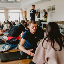 TIME4YOU Sp. z o.o. - Grupa osób siedzi na matach w jasnym pomieszczeniu podczas sesji grupowej. Mężczyzna w niebieskiej koszulce gestykuluje, rozmawiając z kobietą na pierwszym planie.