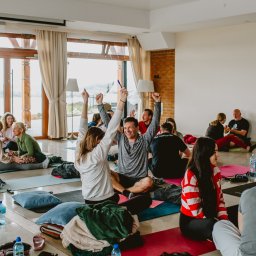 TIME4YOU Sp. z o.o. - Grupa osób siedzi na matach w przestronnym, jasnym pomieszczeniu, z podniesionymi rękami, prawdopodobnie podczas warsztatów lub sesji rozwojowej.