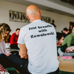 TIME4YOU Sp. z o.o. - Uczestnicy siedzą na podłodze podczas warsztatów oddechowych z instruktorem Kowalewskim, widocznym od tyłu, w białej koszulce z napisem.