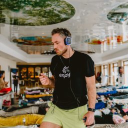 TIME4YOU Sp. z o.o. - Mężczyzna z mikrofonem i słuchawkami prowadzi sesję relaksacyjną. W tle grupa osób leżących na materacach. Sufit z motywem roślinnym. Nietypowe oświetlenie i ustawienie.