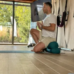 Mężczyzna w jasnej koszulce i beżowych spodenkach siedzi na poduszce do medytacji, wykonując ćwiczenia oddechowe w jasnym pomieszczeniu z dużym oknem i sprzętem do ćwiczeń na ścianie, na ekranie...