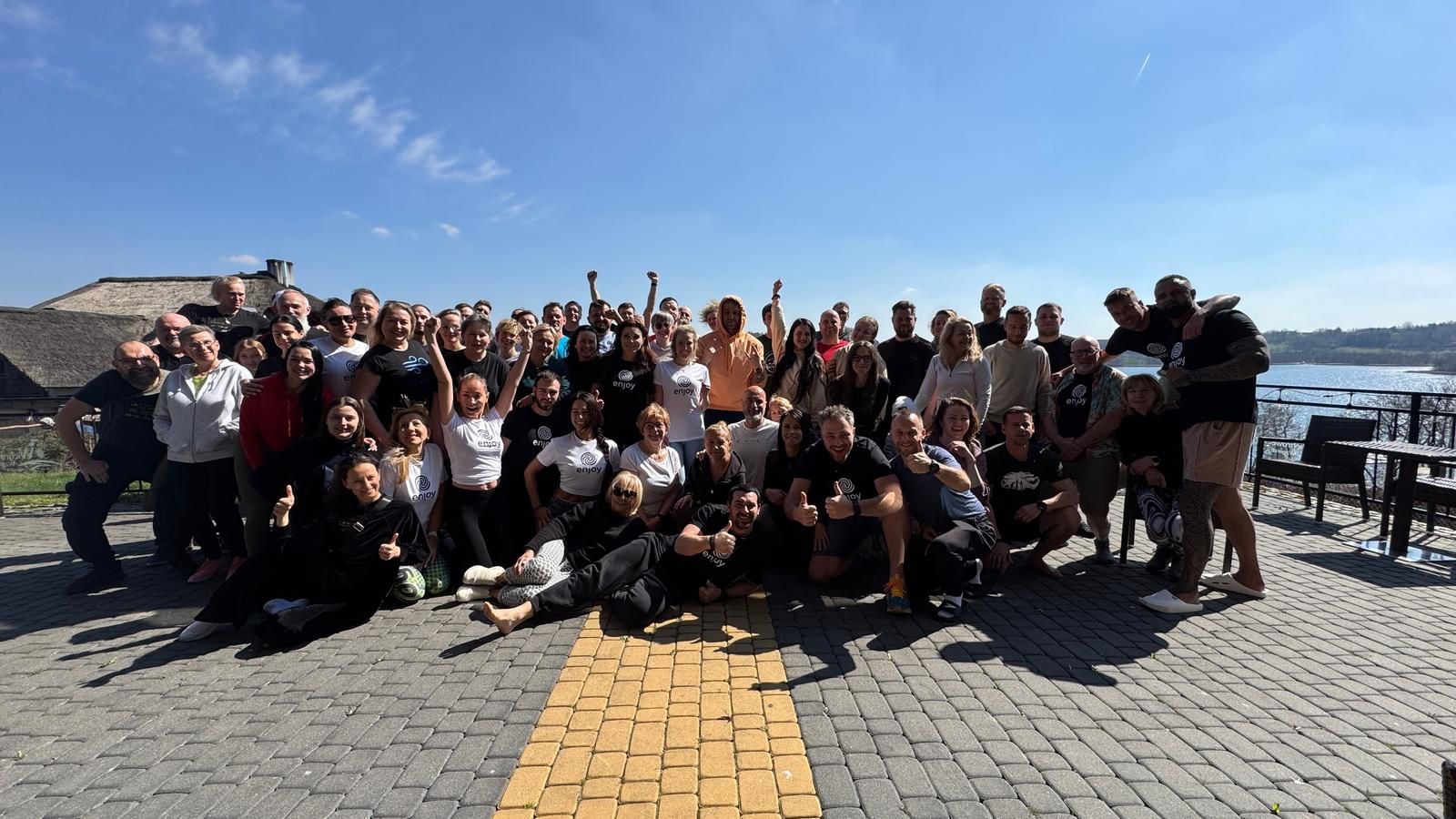 Duża grupa uśmiechniętych osób pozuje do zdjęcia na zewnątrz, część w koszulkach z logo, z widokiem na jezioro i budynek z trzcinowym dachem w tle.