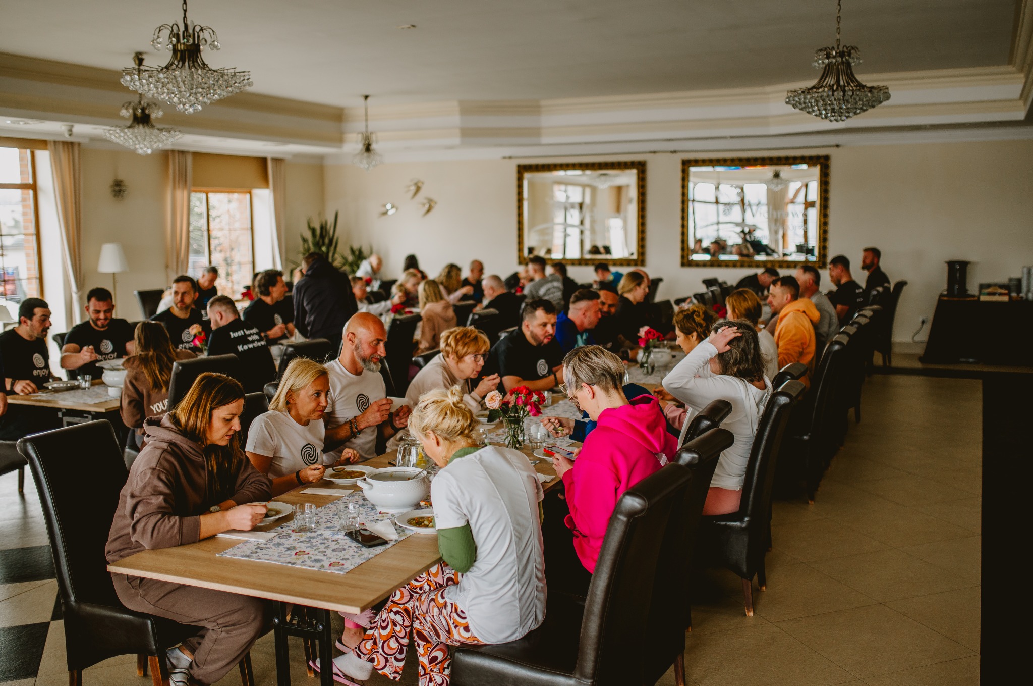 Uczestnicy szkolenia siedzą przy stołach podczas przerwy obiadowej w jasnej sali z lustrami i żyrandolami. Część osób w koszulkach z logo.