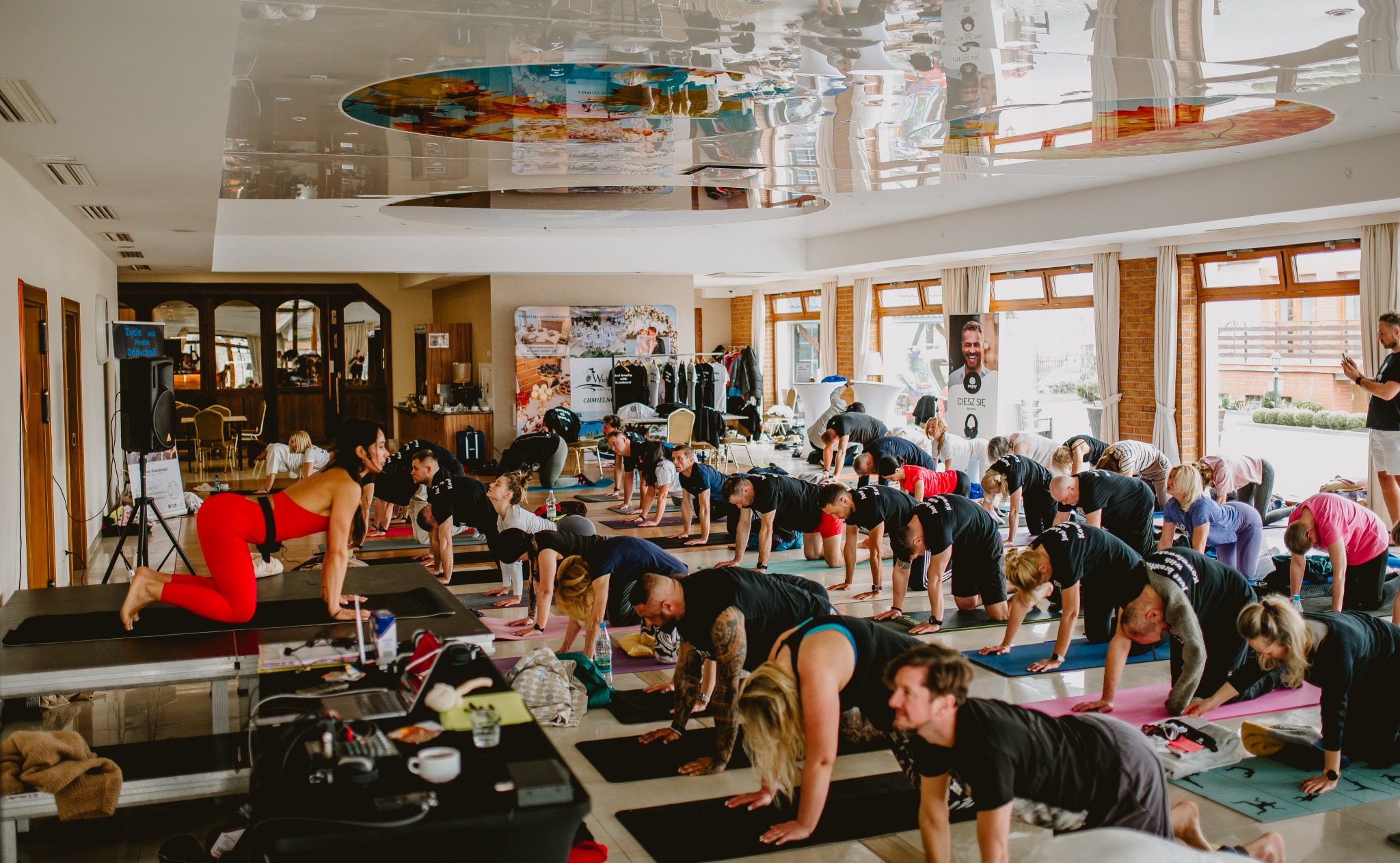 Grupa osób ćwiczy jogę w jasnej sali z lustrzanym sufitem. Uczestnicy w pozycji 'krowy' na matach. Na pierwszym planie stolik z laptopem i reżyserką.