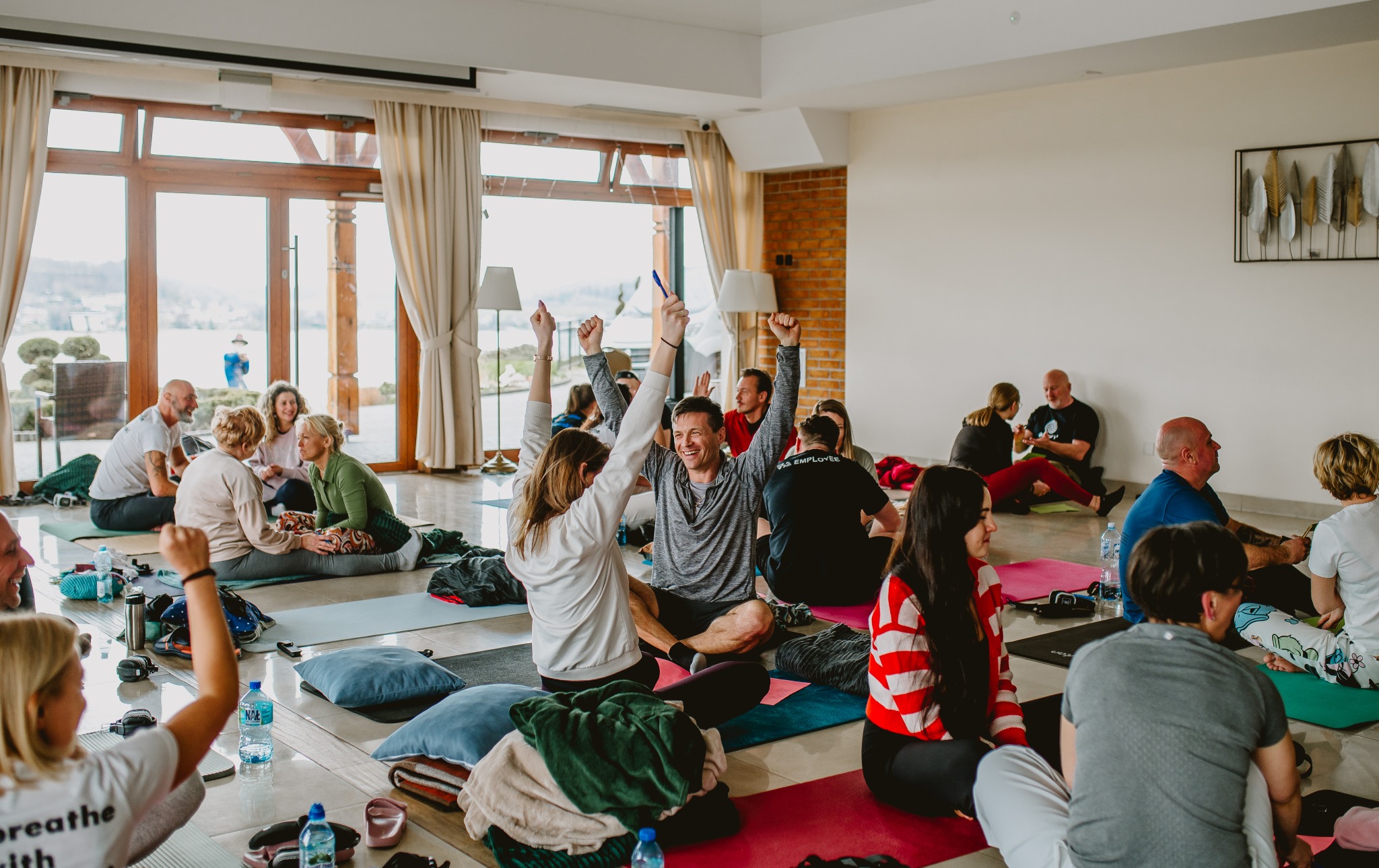 Grupa osób siedzi na matach w przestronnym, jasnym pomieszczeniu, z podniesionymi rękami, prawdopodobnie podczas warsztatów lub sesji rozwojowej.