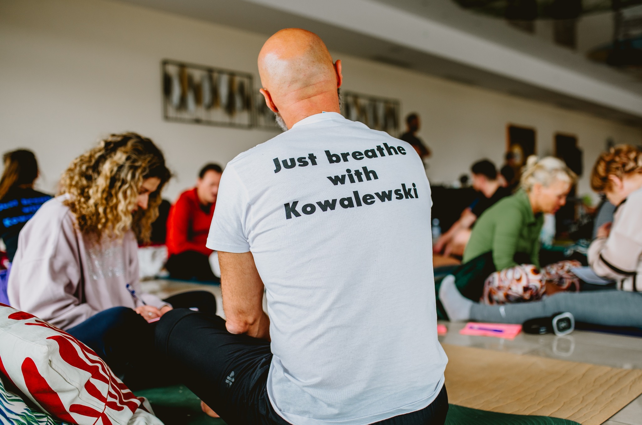 Uczestnicy siedzą na podłodze podczas warsztatów oddechowych z instruktorem Kowalewskim, widocznym od tyłu, w białej koszulce z napisem.