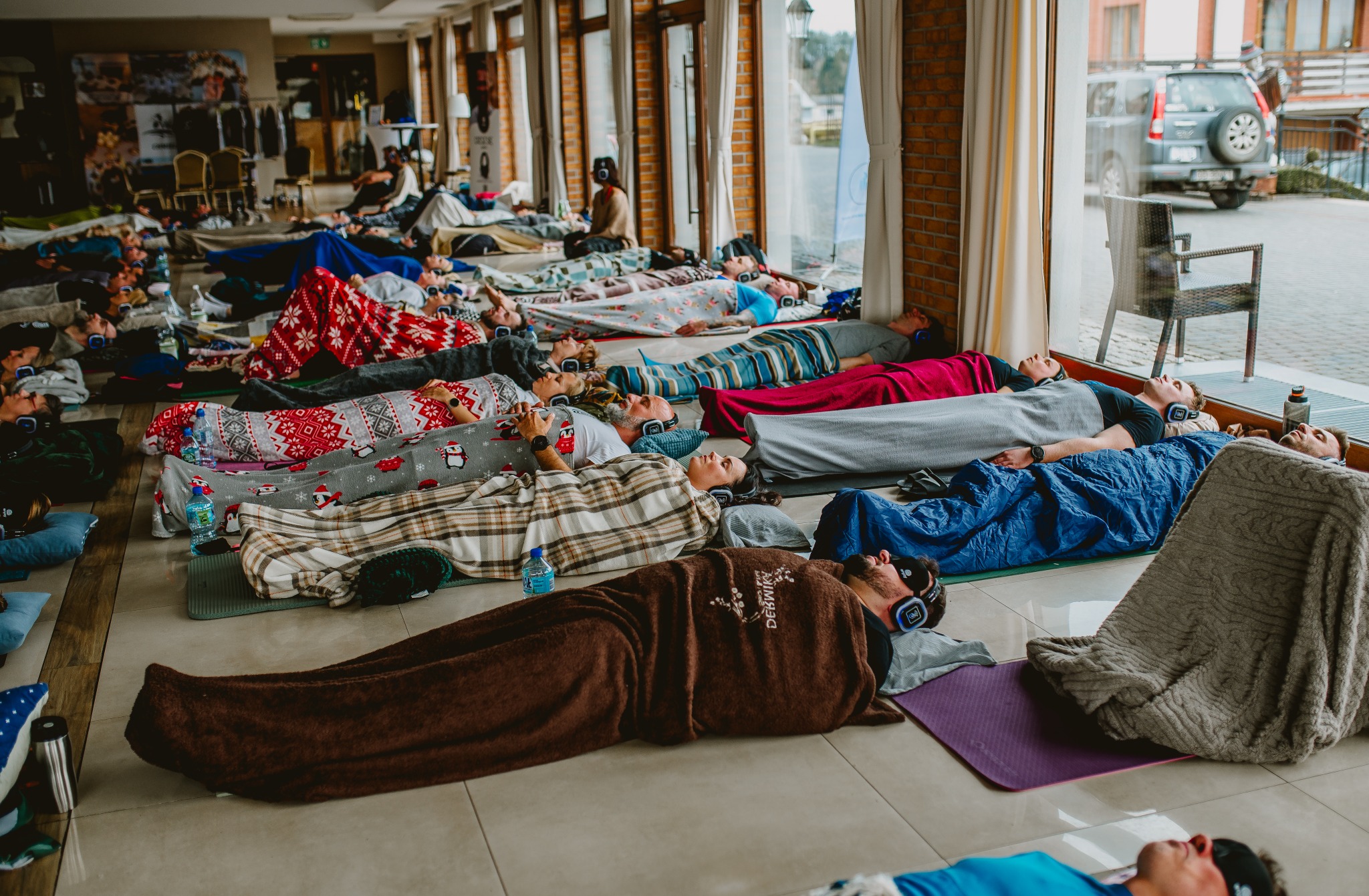 Grupa osób leży na podłodze w słuchawkach podczas sesji relaksacyjnej. Koce, maty do ćwiczeń i butelki wody obok. Widok z góry, jasne wnętrze.
