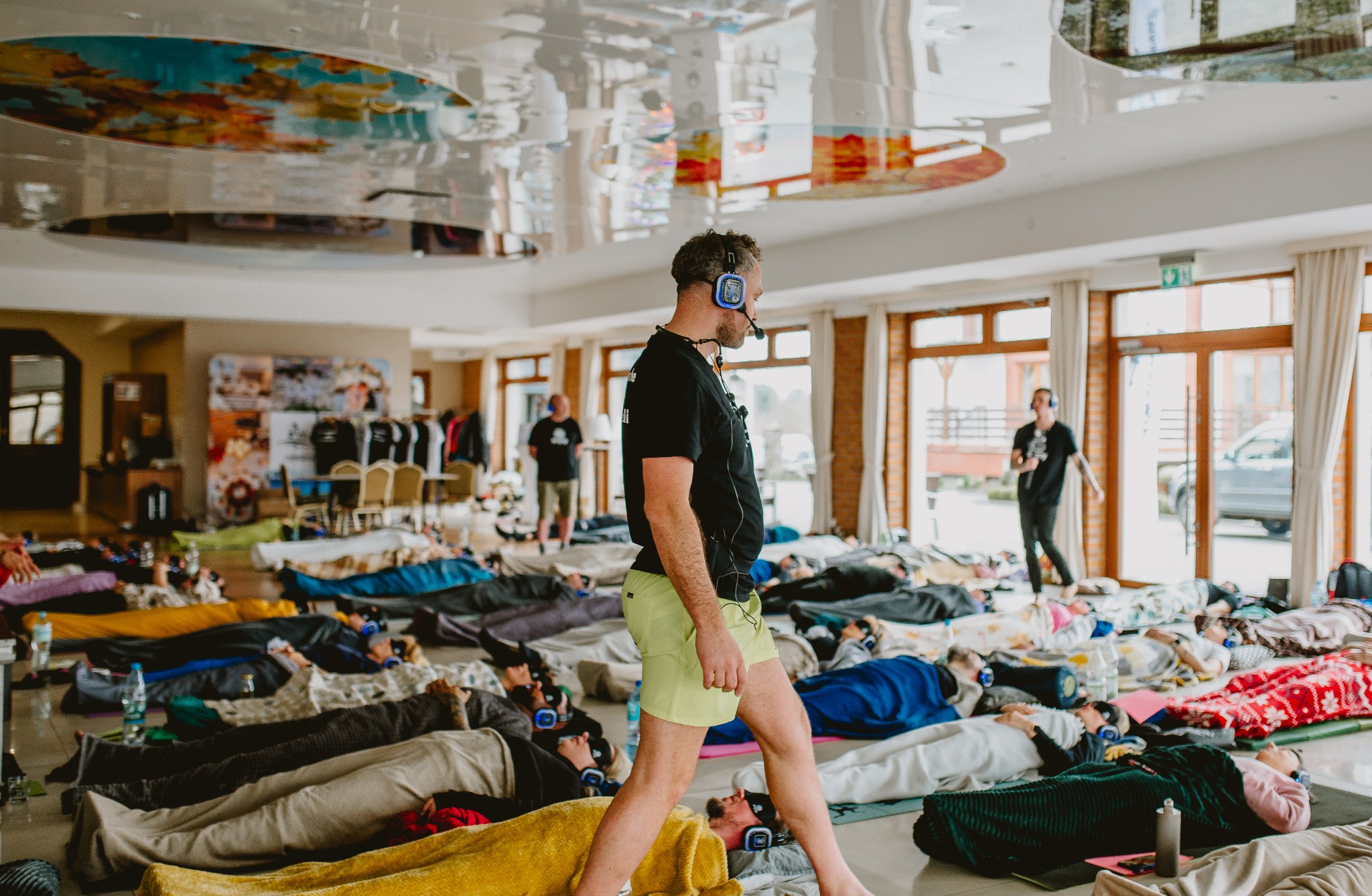 Sala z uczestnikami leżącymi na matach w słuchawkach, prowadzona przez trenera z mikrofonem. Duże okna, jasne wnętrze, odblaskowy sufit. Sesja relaksacyjna.