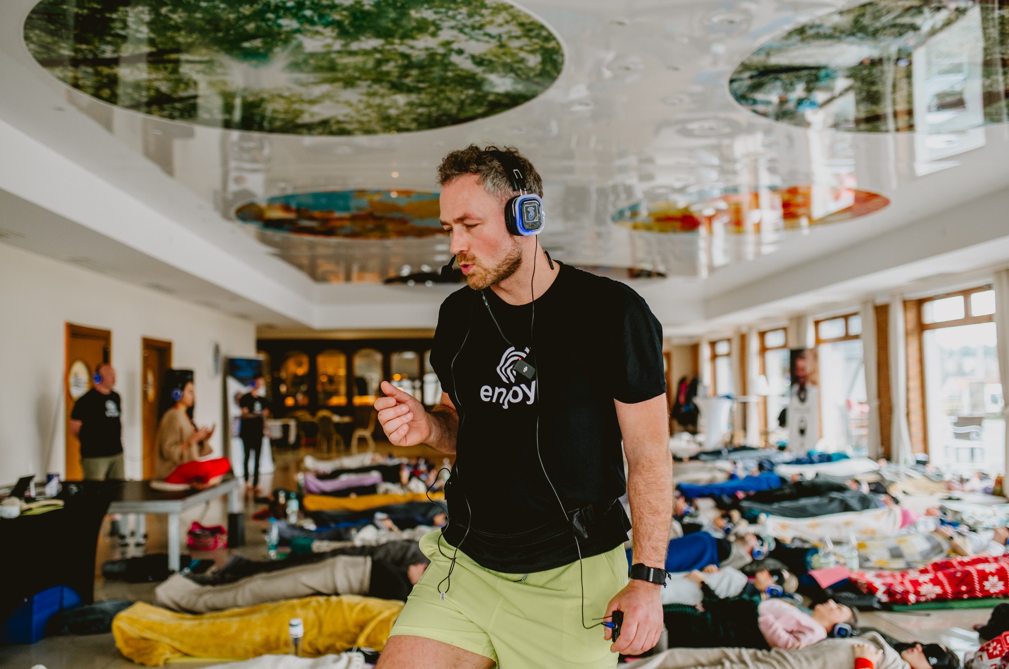 Mężczyzna z mikrofonem i słuchawkami prowadzi sesję relaksacyjną. W tle grupa osób leżących na materacach. Sufit z motywem roślinnym. Nietypowe oświetlenie i ustawienie.