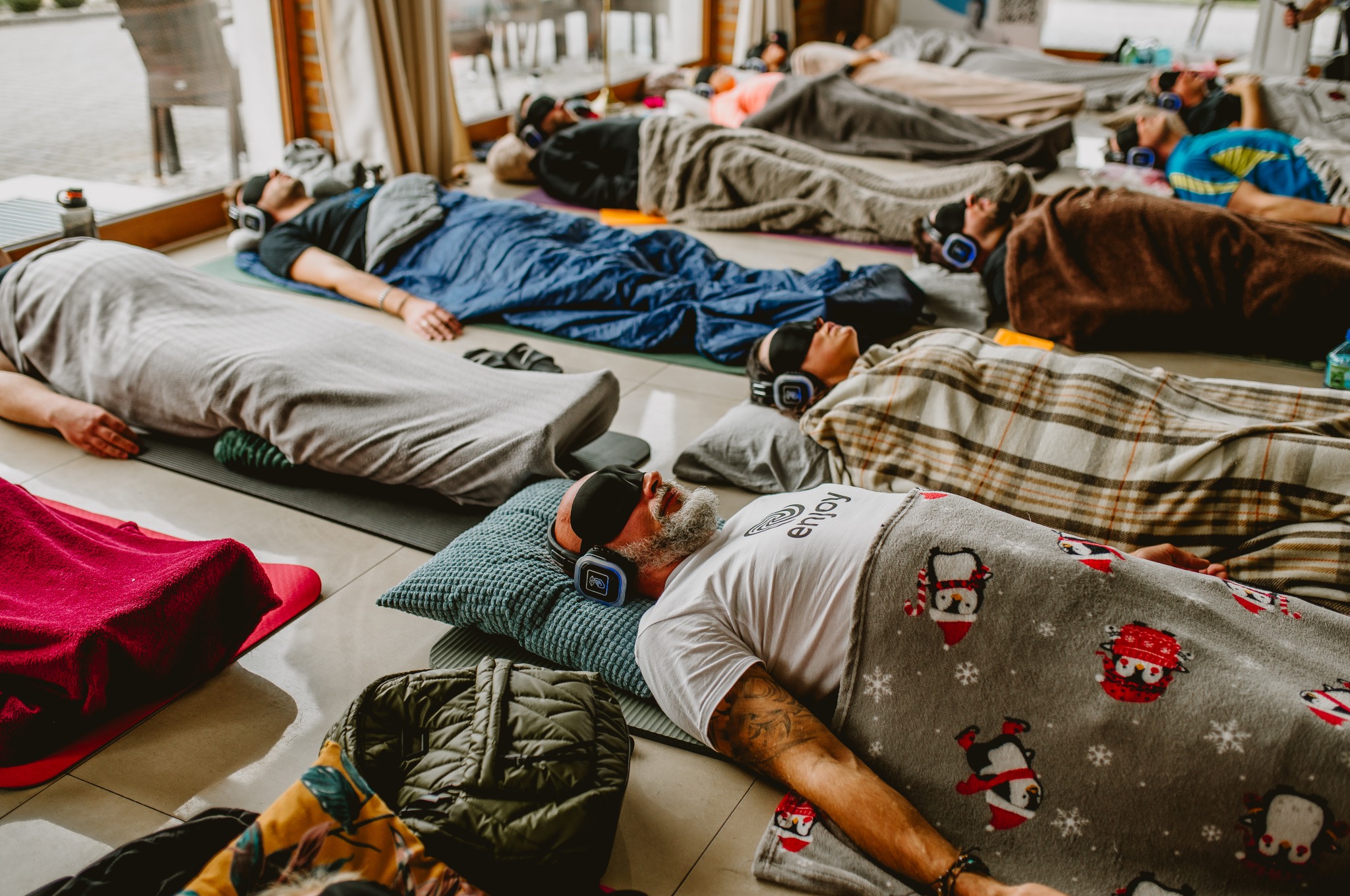 Grupa osób relaksuje się w pozycji leżącej na podłodze, zakryta kocami, z opaskami na oczach i słuchawkami na uszach, podczas sesji relaksacyjnej.